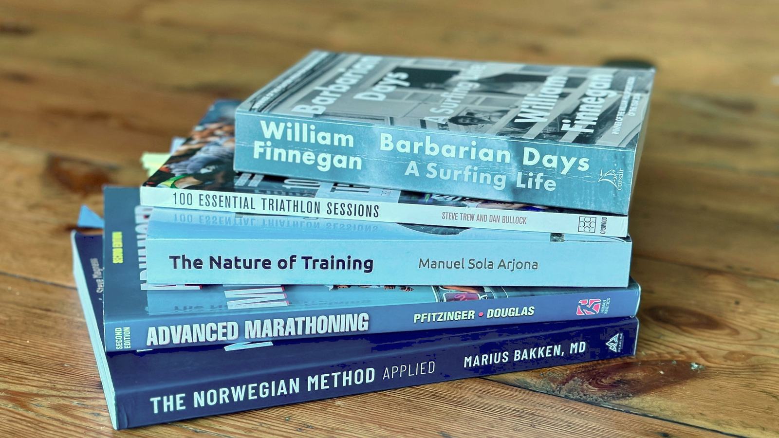 A stack of five well-read books on a wooden table: Barbarian Days, 100 Essential Triathlon Sessions, The Nature of Training, Advanced Marathoning and The Norwegian Method Applied