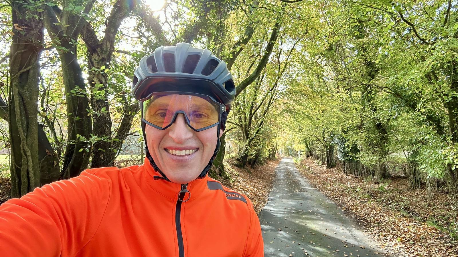 Nick Buckley-Mills smiling in an orange cycling jacket and helmet on a quiet, leaf-covered lane under autumn trees